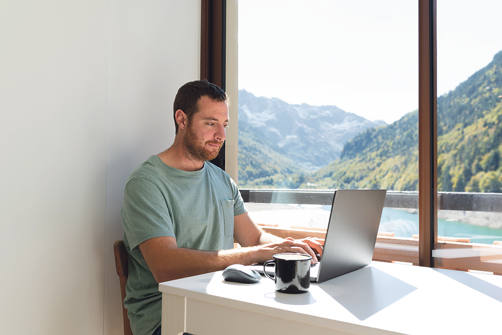 Ein Mann sitzt zu Hause vor einem Panoramafenster am Laptop.